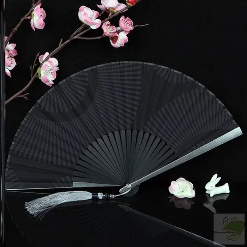 A black titanium alloy folding hand fan with a white tassel is displayed against a dark background.
