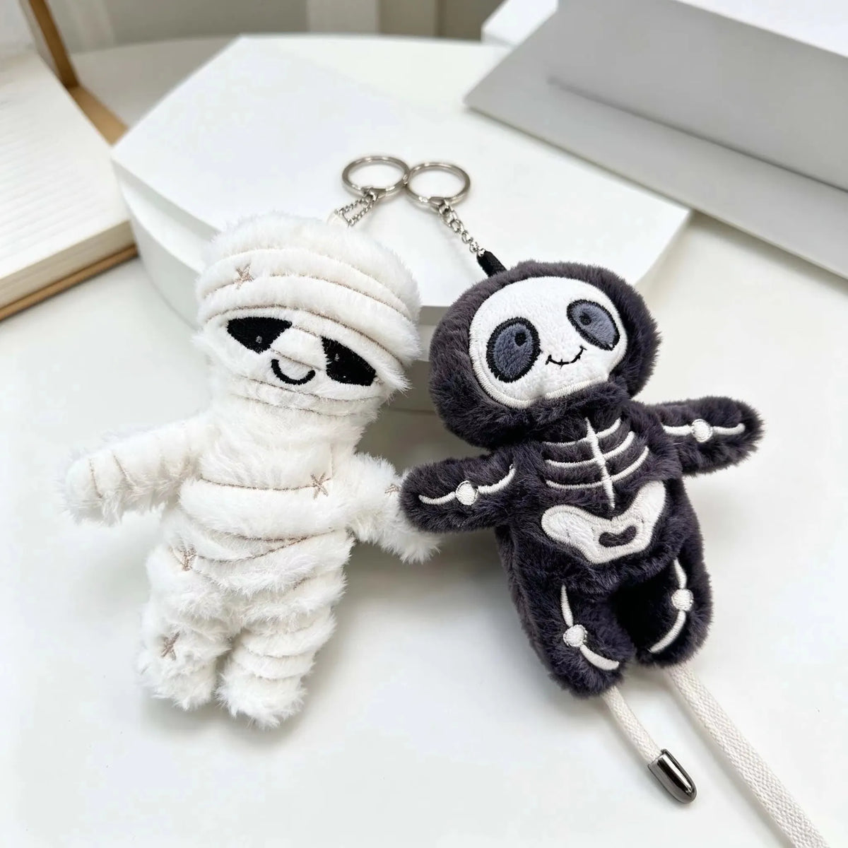 A white mummy skeleton plush keychain with a black skeleton design and a silver chain.