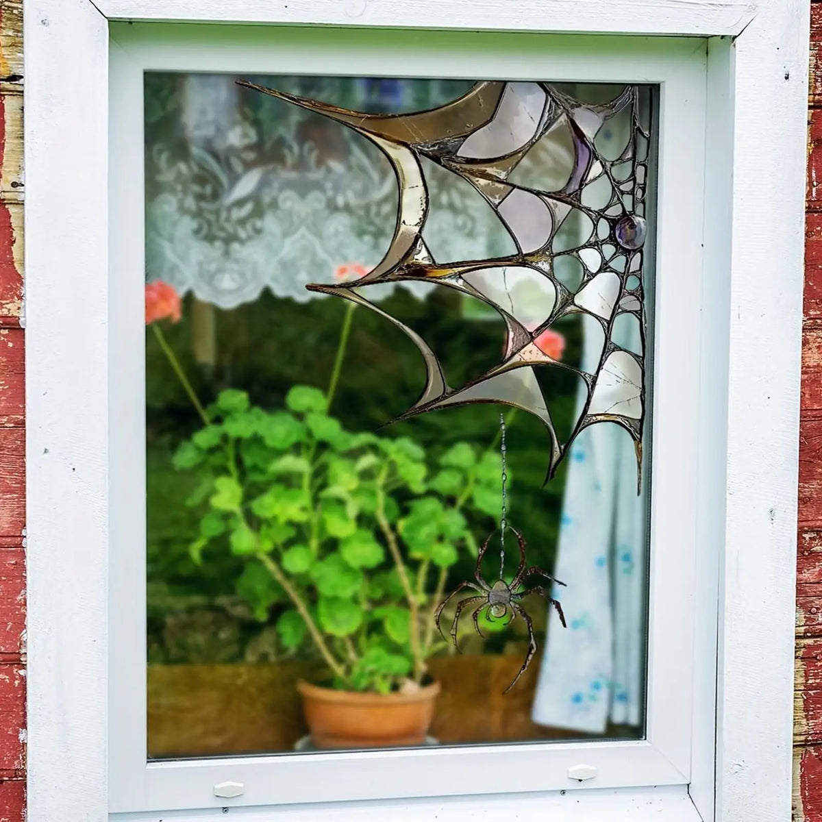 A decorative window decal shaped like a spider web with a small spider hanging in front of greenery.