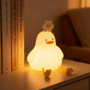 A glowing white duck LED night light with a flower on its head resting on a wooden surface next to books.
