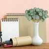 A vase holds artificial eucalyptus stems next to a spiral notebook and an open pencil case on a wooden surface.