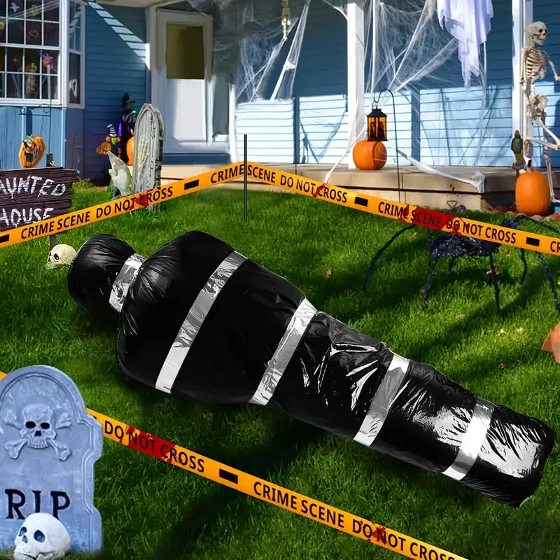 A black body bag with white stripes lies on green grass next to yellow crime scene tape and Halloween props.