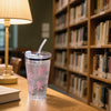 A clear plastic cup with pink bows, a straw, and lid sits on a wooden table in front of a bookshelf.
