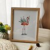 A framed watercolor illustration of a flamingo in a wooden frame on a light-colored surface.