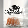 A black graduation cap topper with congratulations text stands above a cake dusted with powdered sugar.