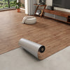 A roll of 3D textured wood grain self-adhesive flooring film placed on a light gray floor next to wooden furniture.