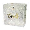 A clear acrylic gift box filled with silver glitter and featuring a gold script word card