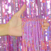 A hand holds a set of metallic pink and purple foil tinsel fringe streamers against a sparkly backdrop.