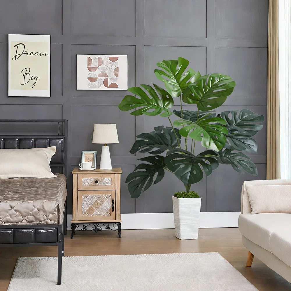 A large artificial palm tree in a white planter sits on a wooden nightstand next to a dark upholstered bed.
