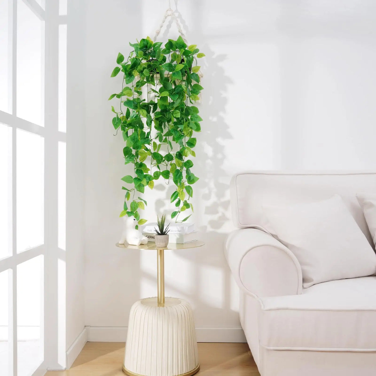 Artificial Pothos hanging vines in a white pot displayed on a glass stand next to a light-colored sofa.