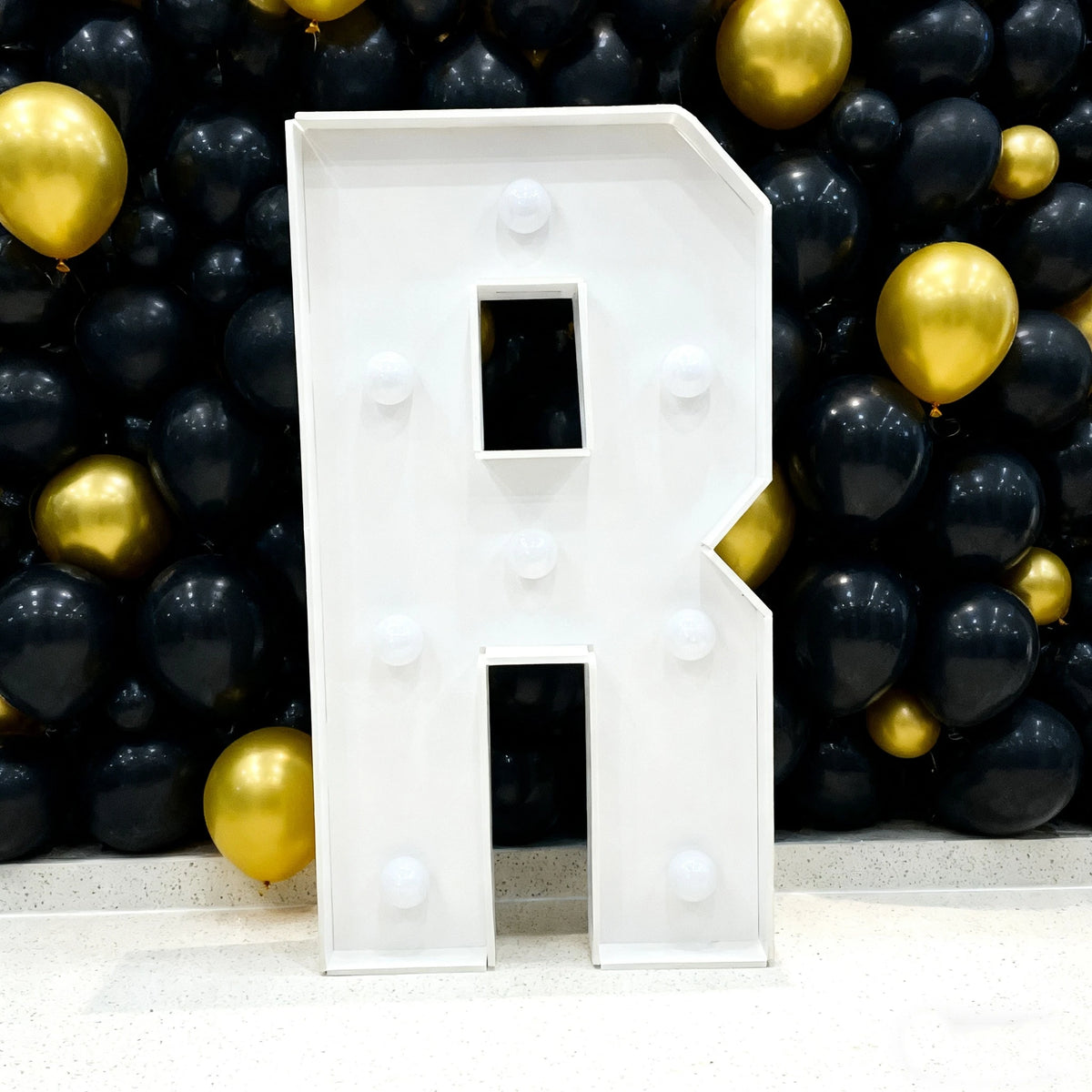 A large white marquee letter R made of acrylic is displayed against a backdrop of black and gold balloons.