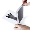 A handprint and footprint ink pad kit featuring a black handprint on white paper, held by a person.