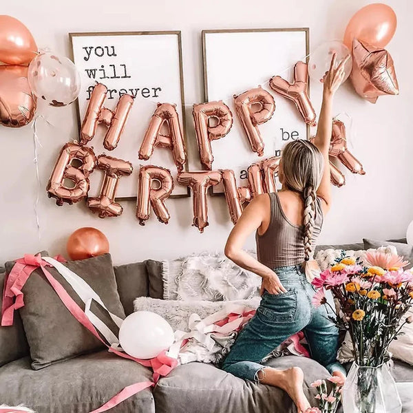 A collection of rose gold foil letter balloons spelling out Happy Birthday arranged on a white wall.