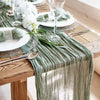 A rustic wooden table set with a sage green semi-sheer gauze table runner, white plates, and greenery.
