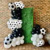 A balloon garland arch featuring black and white soccer balls against a green hedge backdrop.