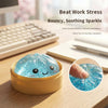 A glowing blue squishy toy shaped like a dumpling resting in a yellow dish on a wooden desk.