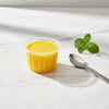 A clear disposable dessert cup filled with bright yellow juice or puree sits on a white marble surface next to a silver spoon