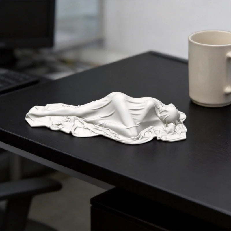 A white 3D printed sculpture of a draped, reclining female figure resting on a dark desk.