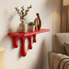 A red drip floating shelf displaying potted greenery and a wooden sculpture on a white wall.