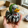 A hand holds a gray and black striped resin figurine of a duck with green feathers and an orange beak.