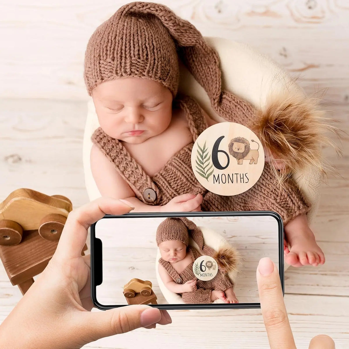 A baby wearing a knitted hat is photographed by a hand holding a smartphone, showing a personalized keepsake sign.