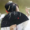 A person holds an open black folding hand fan with white decorative elements in a traditional setting.