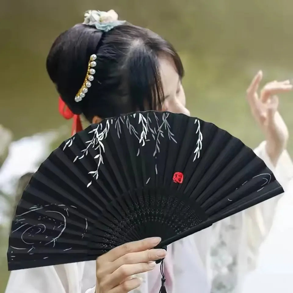 A person holds an open black folding hand fan with white decorative elements in a traditional setting.