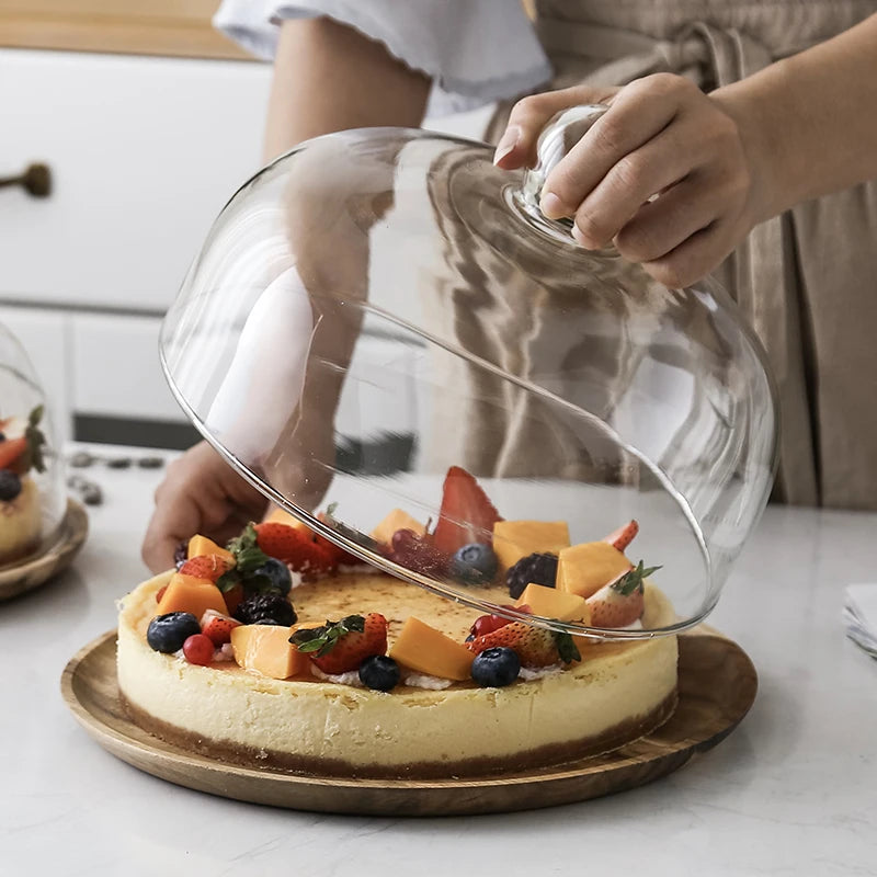A round cake is displayed on a wooden tray with a glass dome cover, being held by a person.