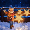 A child wearing a knitted hat and plaid coat holds up several glowing star-shaped pendant lights in a snowy outdoor setting.