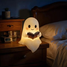 A cute ghost night light shaped like a glowing figure reading a book on a wooden bedside table.