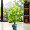 A vibrant artificial floor plant with large green leaves sits in a blue patterned pot on a modern floor.