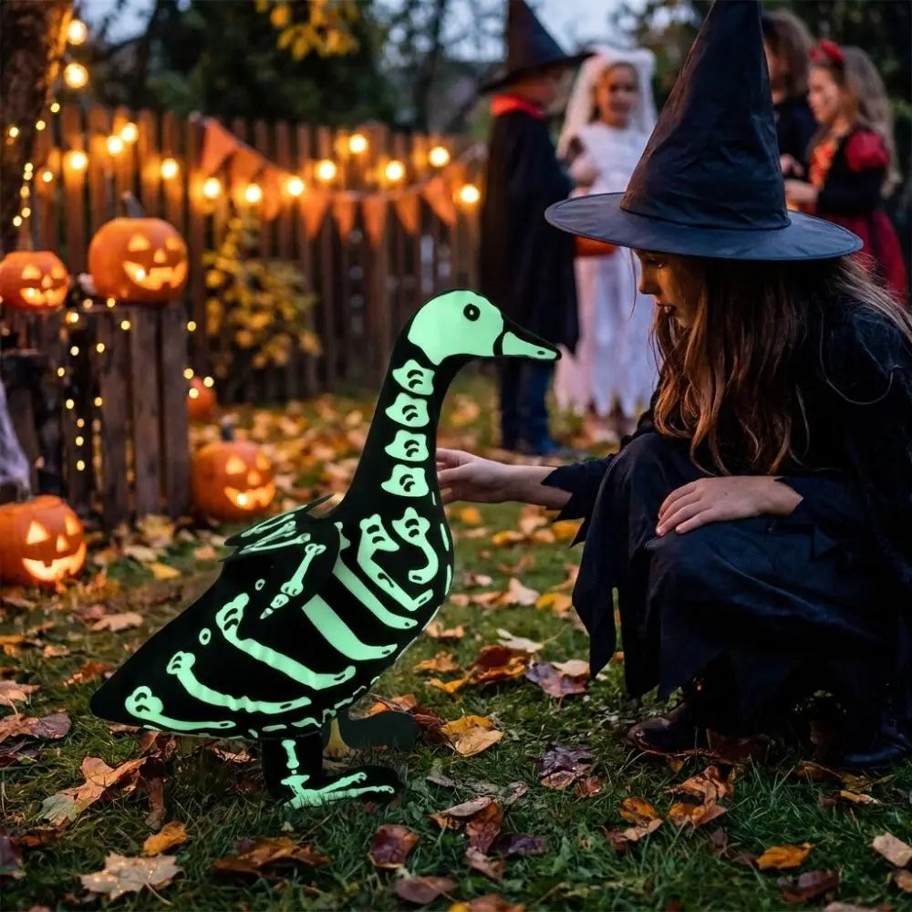 A woman in a witch hat is interacting with a glowing green skeleton goose lawn decor in an autumn outdoor setting.