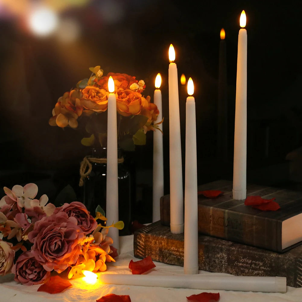 A collection of white taper candles lit in a dark setting next to floral arrangements and stacked books.