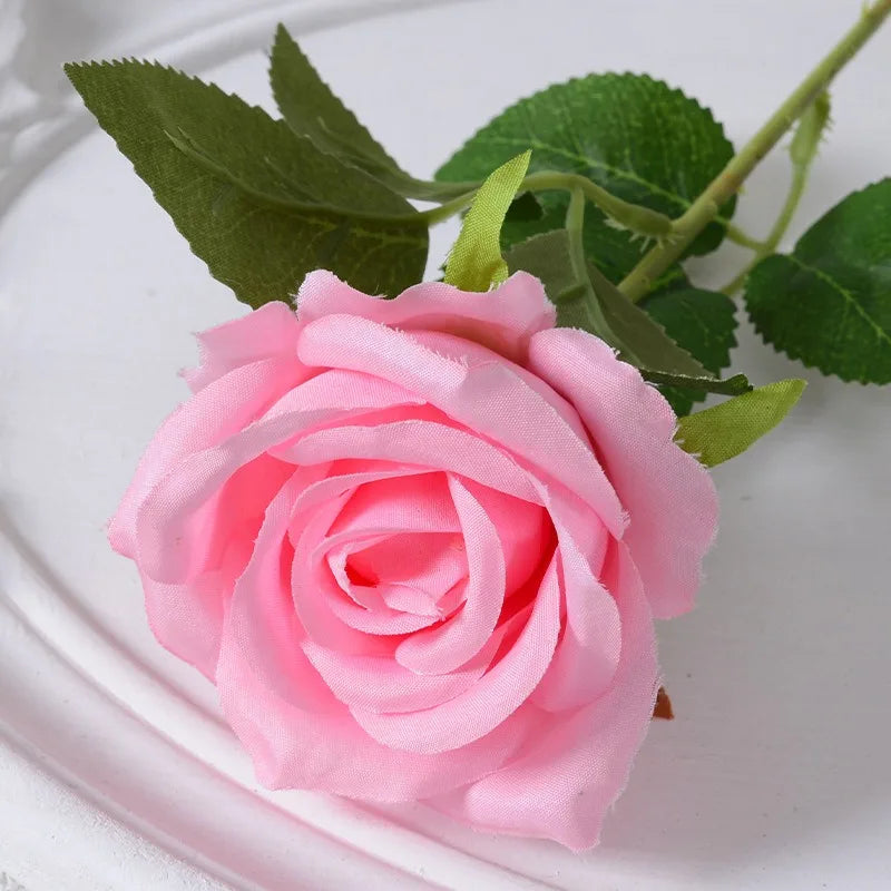 A single pink velvet artificial rose with green leaves on a white plate.