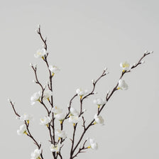 Una rama de flores blancas artificiales de ciruelo sobre un tallo oscuro contra un fondo gris claro.