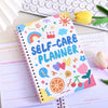 A colorful self-care planner notebook with stickers and pages is shown next to a laptop keyboard.
