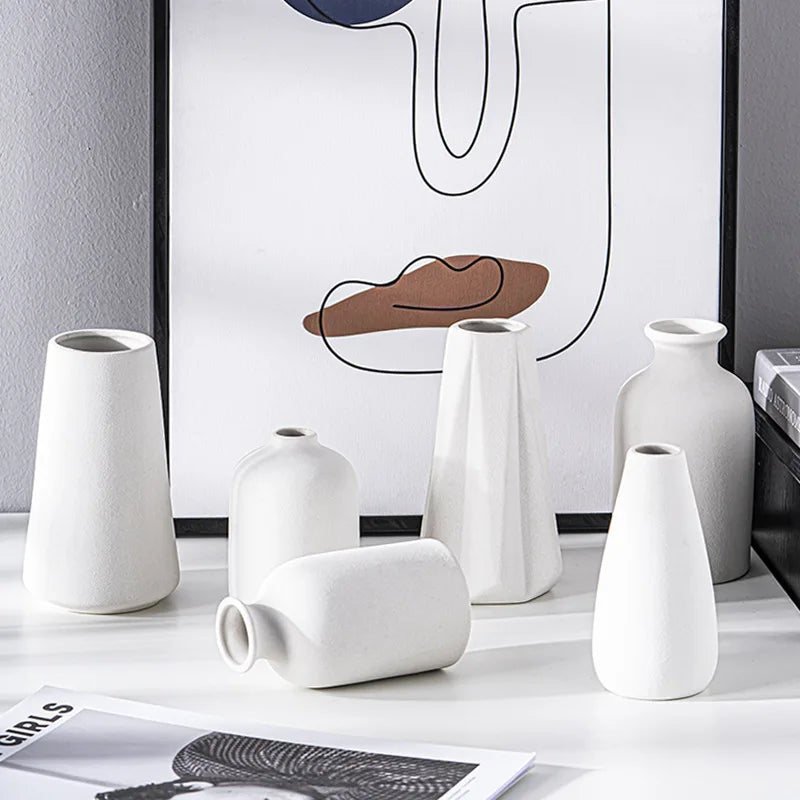 A collection of white ceramic vases and vessels displayed on a white surface against a minimalist artwork.