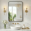 A modern wall mirror with a black metal frame reflects a bathroom interior featuring a white tub and natural light.