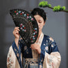 A woman in traditional Japanese attire holds a black decorative folding fan with colorful floral designs.