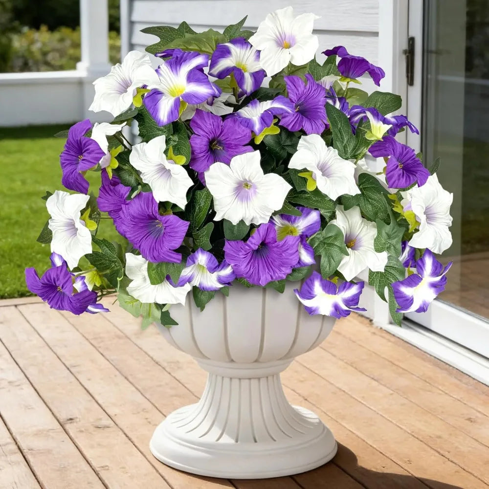 A bouquet of artificial petunia flowers in white and purple are displayed in a white pedestal vase on a wooden deck.