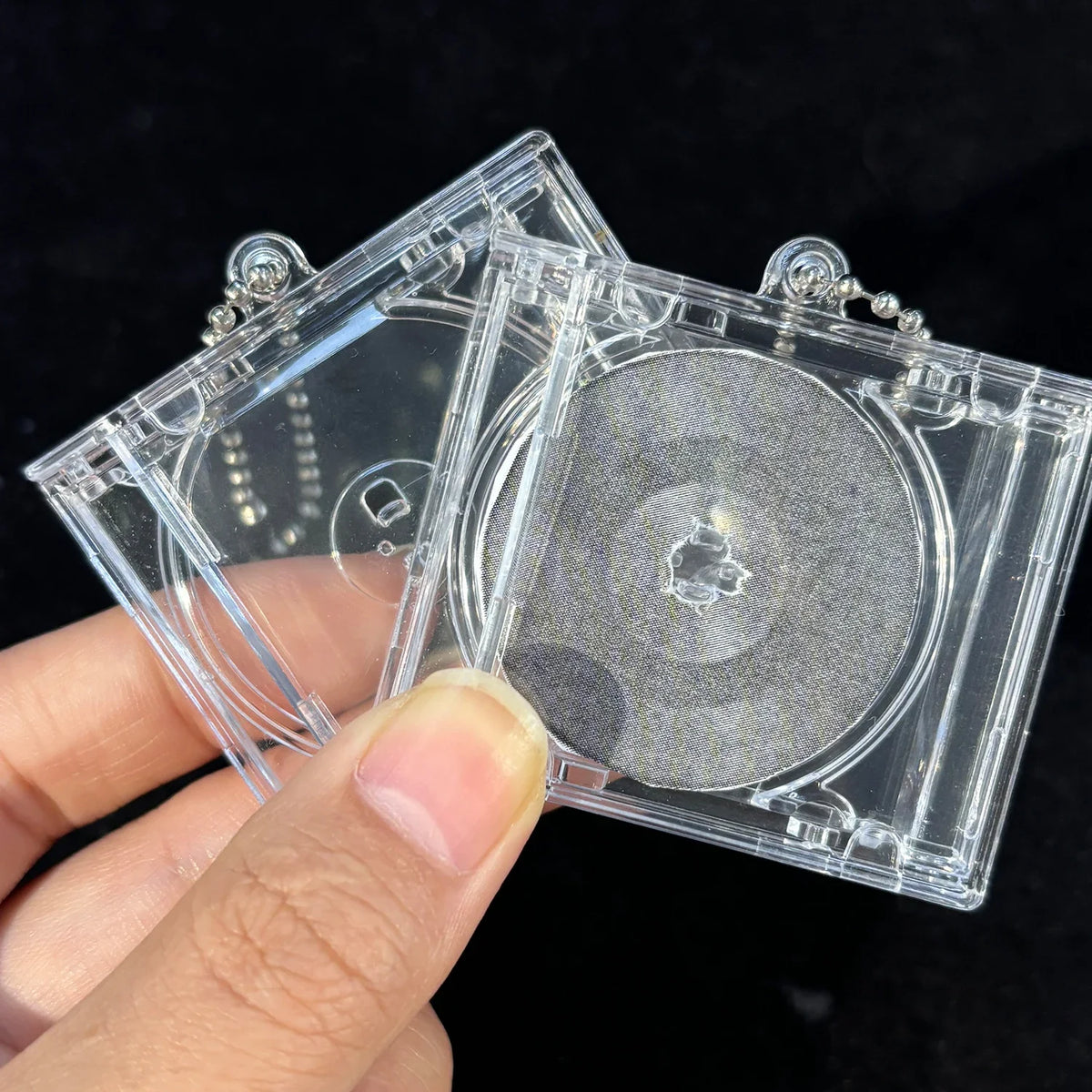 A hand holds a mini transparent CD keychain featuring a silver-colored disc and clear plastic casing.