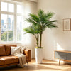 A large artificial areca palm tree in a white pot sits next to a brown sofa by a bright window.