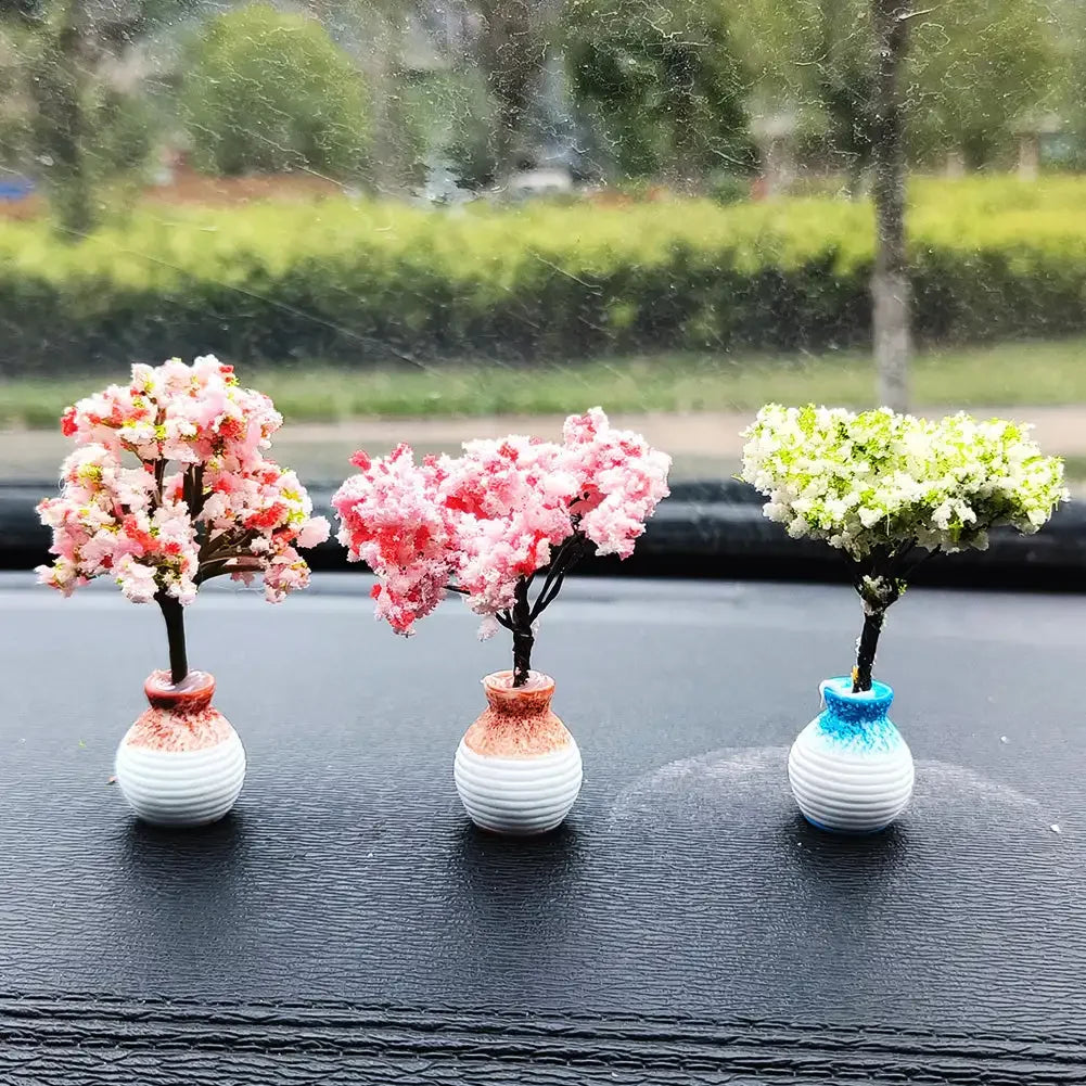 Three small potted trees with pink and white flowers displayed on a black car dashboard.