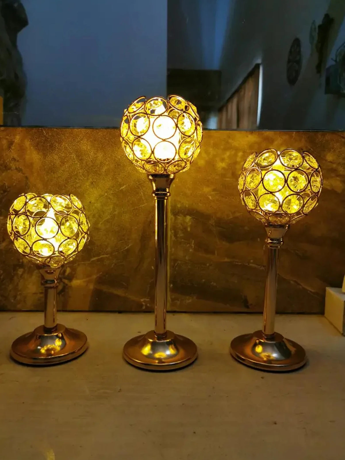 Three golden crystal candle holders with spherical glass bulbs displayed on a light surface.