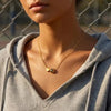 A close-up of a woman wearing a gold heart basketball pendant necklace under a gray hoodie.