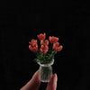 A hand holds a small glass vase filled with vibrant red-orange glass flower sculptures against a dark background.