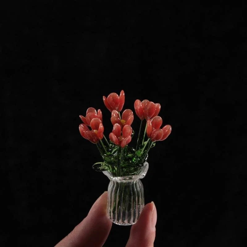A hand holds a small glass vase filled with vibrant red-orange glass flower sculptures against a dark background.