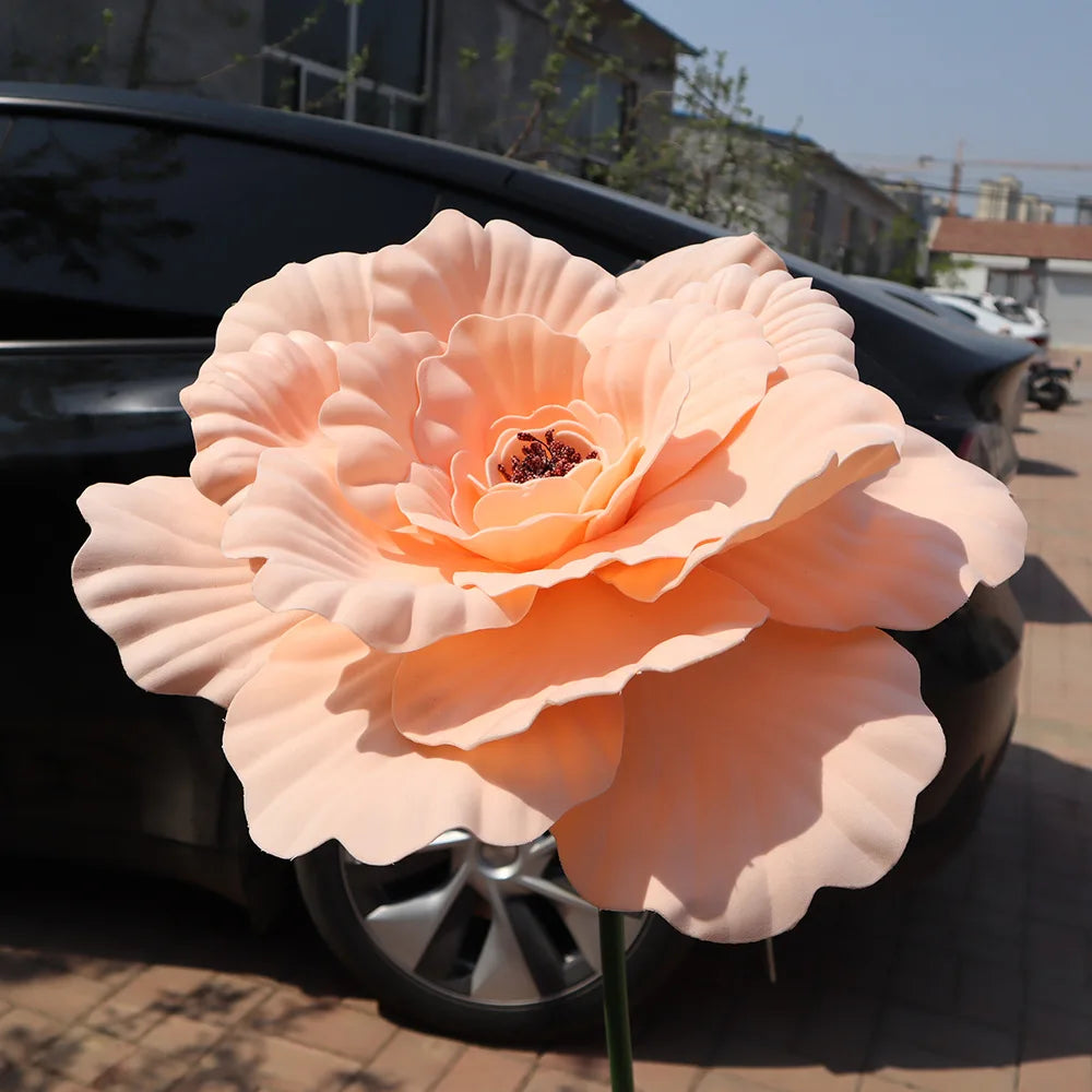 A large, soft peach colored artificial peony flower is displayed in front of a dark car.