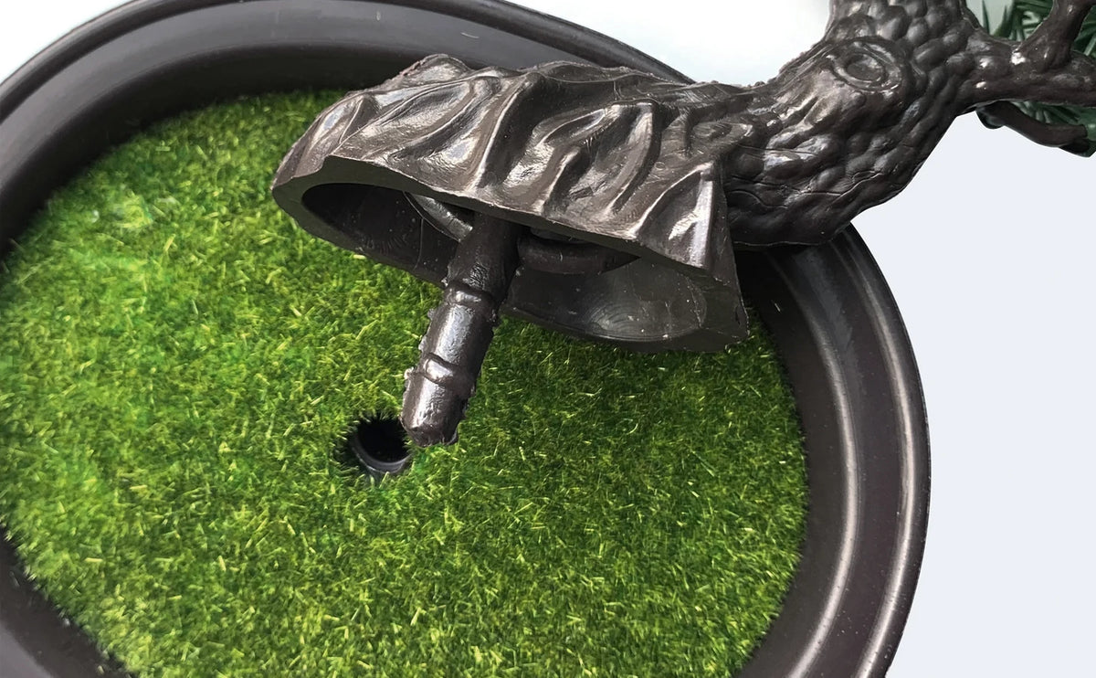 A dark bronze artificial bonsai tree with green foliage is shown emerging from a black pot.