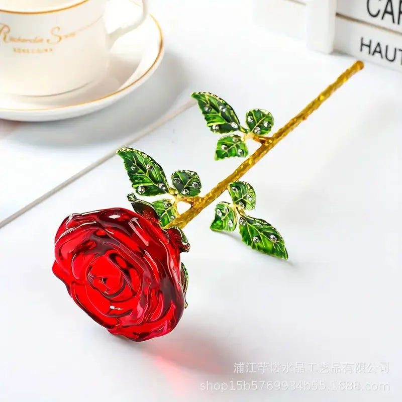 A bright red crystal glass rose flower branch with green leaves resting on a white surface.